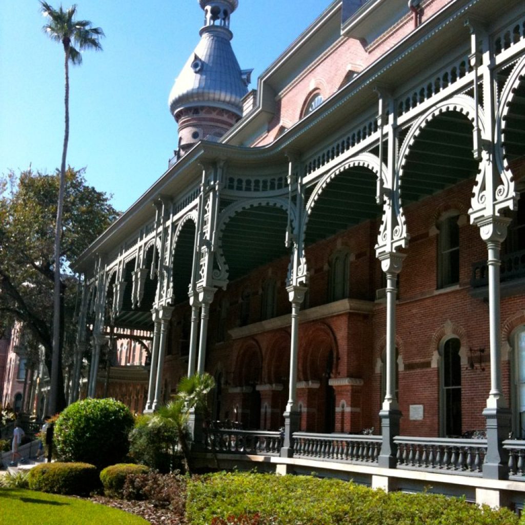 A low angle shot of one of the buildings at The University of Tampa, which is made of red bricks and ornate white columns through out the balcony of the building. In front is a manicured lawns and well-trimmed shrubs. The building is shot during daytime.